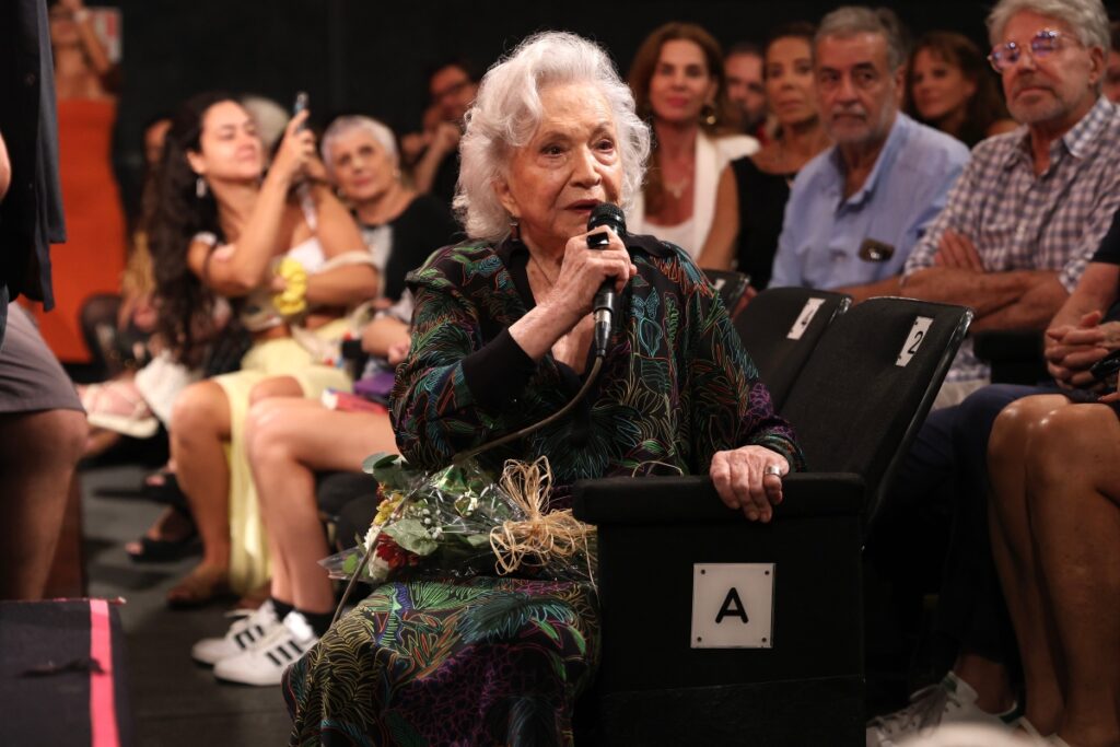 Nathalia Timberg falando ao microfone, sentada